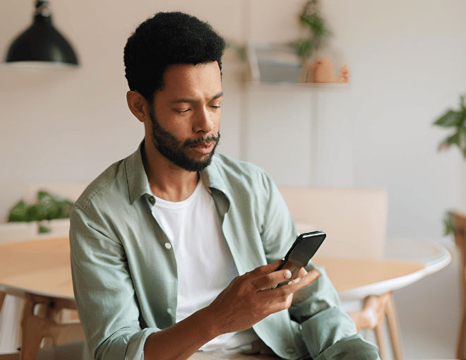 A man in a casual shirt is holding a smartphone, focusing intently on the screen with his thumb poised.